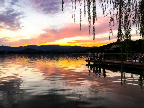 A group of about ten people stand on the end of a small pier on the shore of Beijing’s Beihai Lake. The setting sun beyond the distant hills turns the sky shades of lavender, pink, and egg-yolk yellow, hues that are reflected in the rippling surface of the lake.