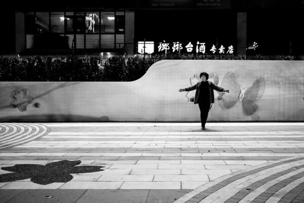 Standing in a public square in front of a butterfly-themed mural, a woman in dark slacks and jacket and a white surgical mask spreads her arms as if preparing to fly.