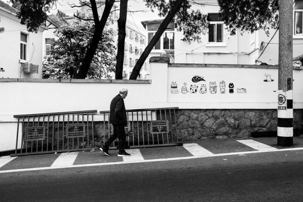Black and white image of a man walking uphill against a backdrop of white multistory buildings and a white wall with a mural of cartoon cats in Qingdao, China.