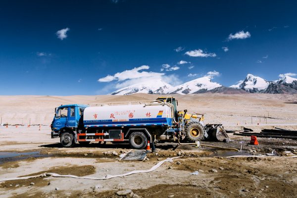 A yellow bulldozer and blue-and-white municipal water tank truck with hoses form a stark contrast with the white sandy soil, brilliant blue sky and distant snow-covered peaks of the Taxkorgan Pass in Taxkorgan Tajik Autonomous County, Kashgar, near the border between Xinjiang and Tajikistan.