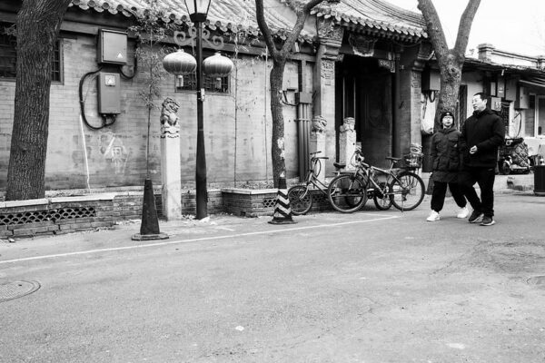 A father and son, dressed warmly in padded jackets, chat as they walk past a traditional brick-faced, tile-roofed, Beijing courtyard-style house adorned with lanterns.