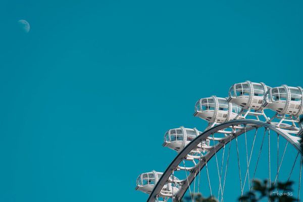 Five white metal capsules of a giant ferris wheel stand out clearly against a brilliant turquoise sky; in the top left corner, the moon is faintly visible. (The Bay Glory, opened in April 2021, is a giant cantilevered observation wheel located at Qianhai Bay, Shenzhen, China.)