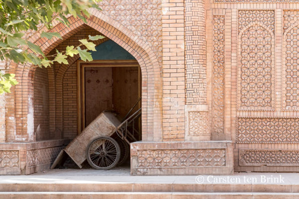 A tilted, two-wheeled wooden cart rests in an arched alcove leading to the doorway of a building in Kashgar Old Town, Xinjiang. The monochrome building of peach-colored brick features many of the familiar geometric patterns found in Islamic architecture.