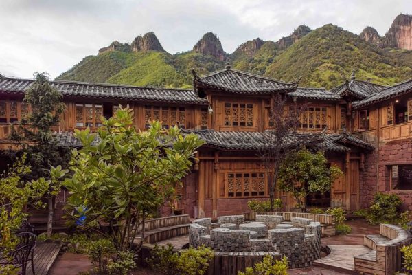 A traditional Yunnanese courtyard home features red stone, intricate woodwork, tile rooftops, lush greenery, and a circular seating area of brick and stone. Behind the house tower the green peaks of the Laojun mountain range, famed for the biodiversity of its flora and fauna.