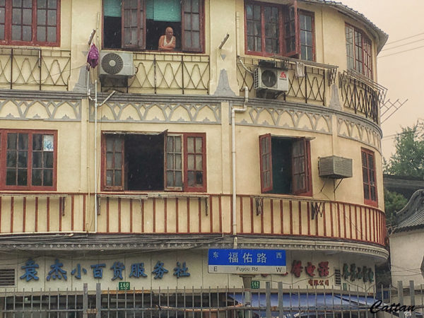 An older man in a white undershirt leans out the top-story window of an old three-story Shanghai building with rounded exterior walls, a weathered yellowed facade, and peeling, red-painted wooden window sills. At street level, a blue-and-white street sign reads, “Fuyou Road.” (“Fuyou” means blessings and protection.)