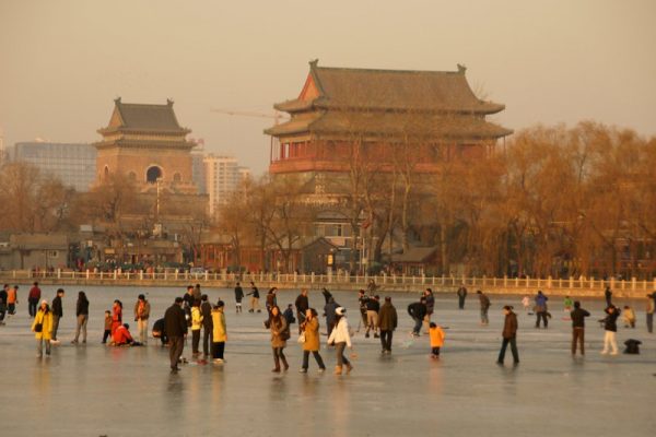 Several dozen warmly dressed skaters enjoy ice-skating on an outdoor lake in Shichahai, Beijing. In the background are several modern high rises and the red columns and tiled rooftops of Beijing’s ancient Drum and Bell Tower.