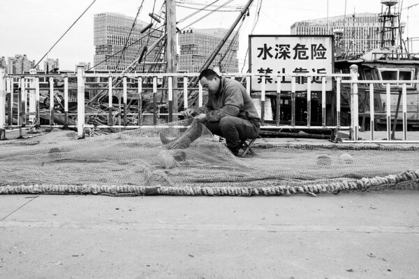 In Qingdao, a port city in China’s Shandong Province, a man in dark jeans, fleece sweatshirt, and sleeve protectors sits on a low stool as he repairs holes in an enormous fishing net. Behind him is a metal railing, a sign warning people to stay away from the railing because the water is deep, and several moored fishing boats; further in the distance are three towering high-rises of light-colored concrete and glass.