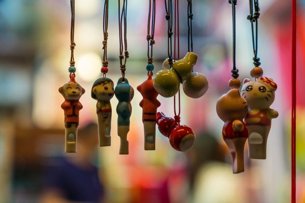 Close-up of eight brightly colored clay whistles in various shapes such as Chinese gourds, and Chinese and Japanese cartoon characters. The whistles, hanging from strings, are clear in the foreground; the background is a blur of bright colors.