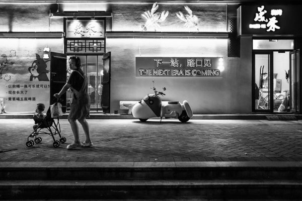 A woman in shorts and sandals walks along a sidewalk, pushing a baby in a stroller. A bilingual neon sign on the facade of a restaurant behind her reads (in all-caps), “THE NEXT ERA IS COMING.”