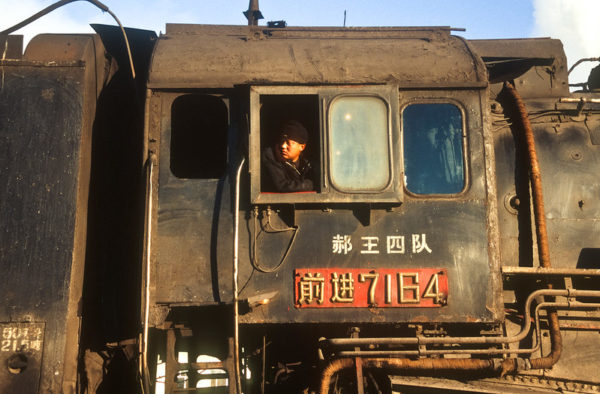 In the engineer’s cabin of a weathered black steam engine, a man in a black jacket and woolen hat rests his shoulder on the sill of an open window and gazes back at the train behind him.