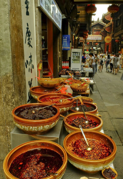 In a narrow alleyway lined with traditional-looking shops and filled with shoppers, an outdoor display features various kinds of chili peppers, chili oils, and chili sauces in large earthenware bowls.