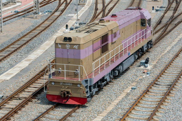 A lone train engine car—painted in bright shades of red, tan, and lavender—sits in a railway yard in Dalian, China, amidst a maze of train tracks.