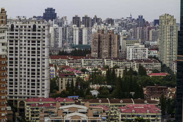 A forest of white, off-white, peach and glass skyscrapers in Shanghai, interspersed with several stands of trees and lower buildings with red rooftops.