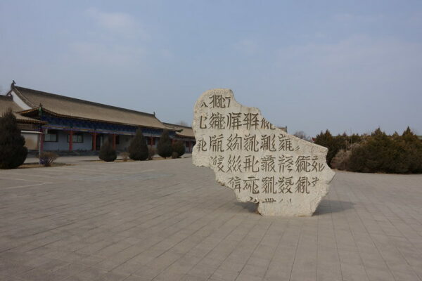 An enormous partial stele displaying Tangut script, a now-extinct language, occupies the center of a vast paved square lined with trees and a long, low building with red pillars and tiled rooftops.