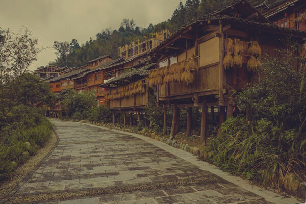 Along the right side of a curved path paved with rectangular gray stones are several rows of small, neat, traditional wooden houses on stilts, topped with tile rooftops and adorned with bamboo or rattan window shades. In the back row, further up the hillside, the frameworks of two new houses in the same style are visible.