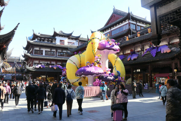 Shanghai shoppers fill a courtyard surrounded by two- and three-story traditional Chinese buildings adorned with intricate woodwork, peaked eaves, and tiled rooftops. In the center of the courtyard is a large inflatable sculpture of a yellow and white snake and purple and lavender toadstools to commemorate the 2025 “Year of the Snake” Chinese Lunar New Year holiday.
