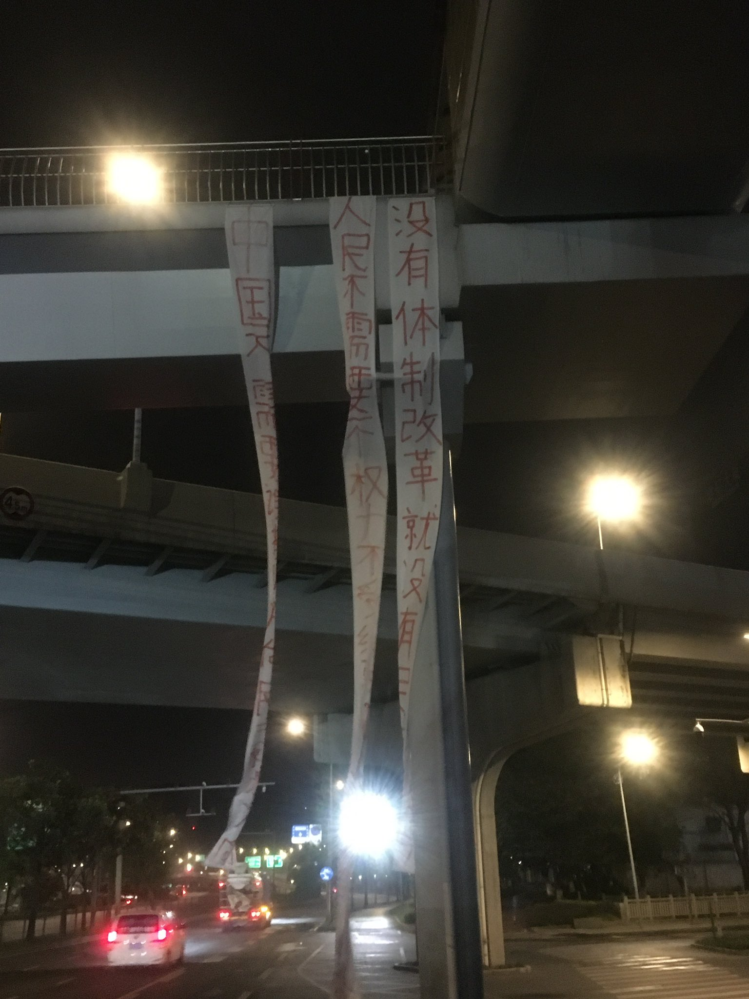 Three long white banners hang from an overpass, twisting in the wind. The sky is still dark, the streetlights are on, and the taillights of two vehicles—a car and a truck—glow red as they pass by on the left. Also at left, several illuminated traffic signs (in blue and green, respectively) are visible in the distance.